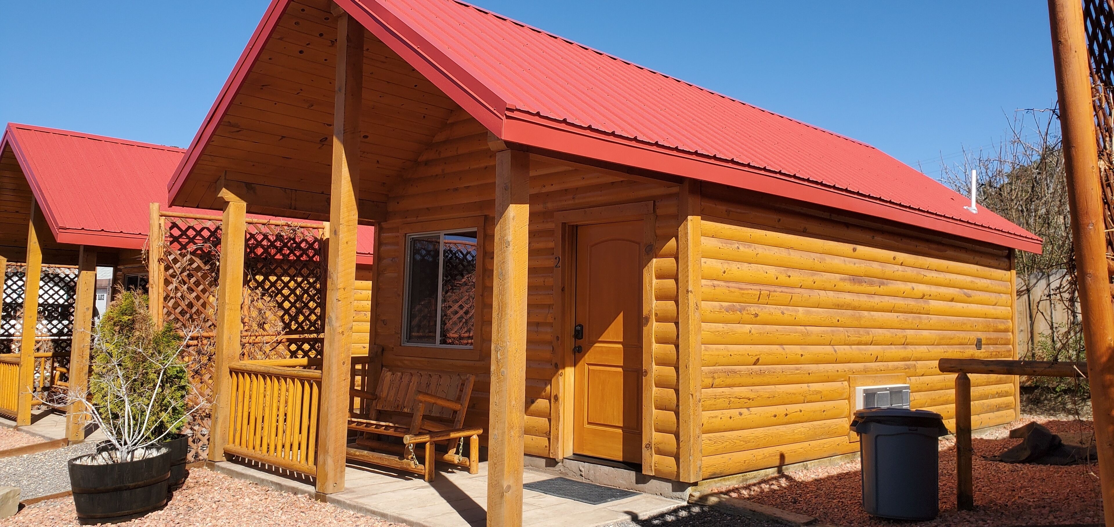 A wooden cabin with a red roof, a porch with a bench, and a small window.
