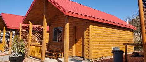 A wooden cabin with a red roof, a porch with a bench, and a small window.