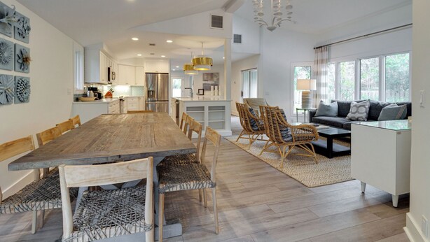 A spacious living area with a large wooden dining table, wicker chairs, a sofa, and a coffee table. There is a kitchen in the background with white cabinets and a hanging light fixture. - 9 Baynard Cove Road at The Sea Pines Resort (Hilton Head Island)
