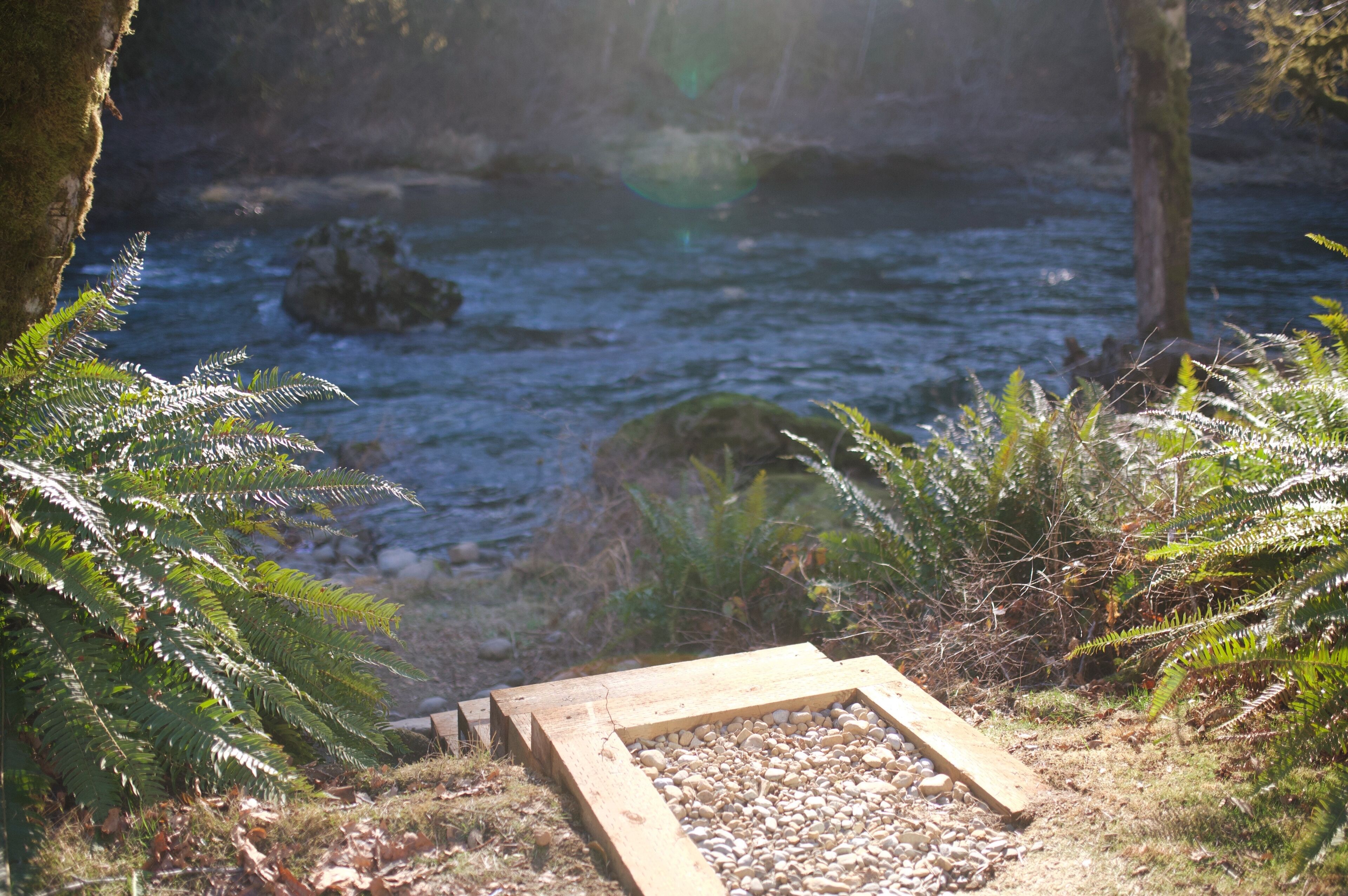Une terrasse en bois bordée de galets, surplombant une rivière et entourée de fougères et de rochers.