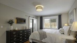 Ein Schlafzimmer mit Bett, Kommode, Nachttisch, Lampe, Fenster mit Vorhängen und einer Deckenleuchte.