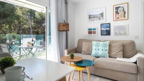 A modern living room with a sofa, a coffee table, and a dining area with a table and chairs. There are framed artworks on the wall and a view of the outdoors through large windows. - Sunny apartment 1B with balcony & pool in canyamel (Capdepera)