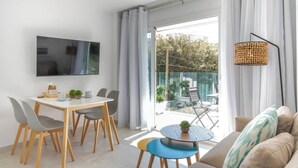 A modern living room with a sofa, a dining table with chairs, a TV mounted on the wall, and a balcony with a view of the outdoors. - Sunny apartment 1B with balcony & pool in canyamel (Capdepera)