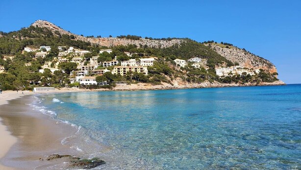 A coastal beach with clear water, a rocky foreground, and a hillside with buildings. - Sunny apartment 1B with balcony & pool in canyamel (Capdepera)
