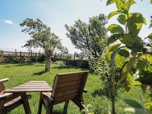 Una panchina e un tavolo di legno in una zona verdeggiante all'aperto, con alberi e una struttura in pietra sullo sfondo.