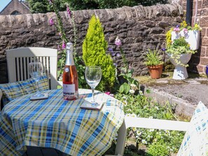 A table set outdoors with a bottle and glass, surrounded by plants and a stone wall. - The Netty (Buxton)