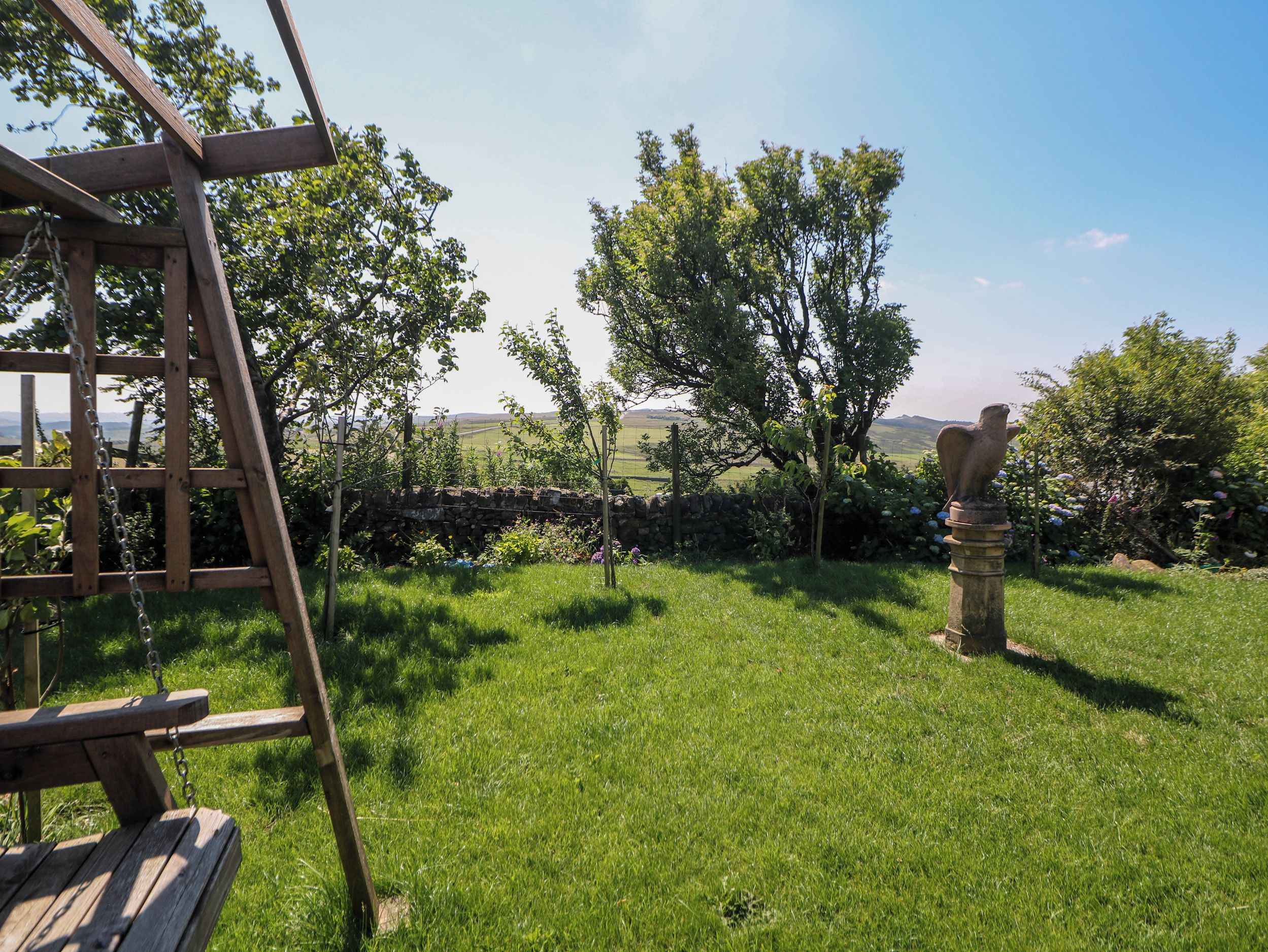 A wooden swing set and a stone wall in a grassy garden with trees and a clear sky.