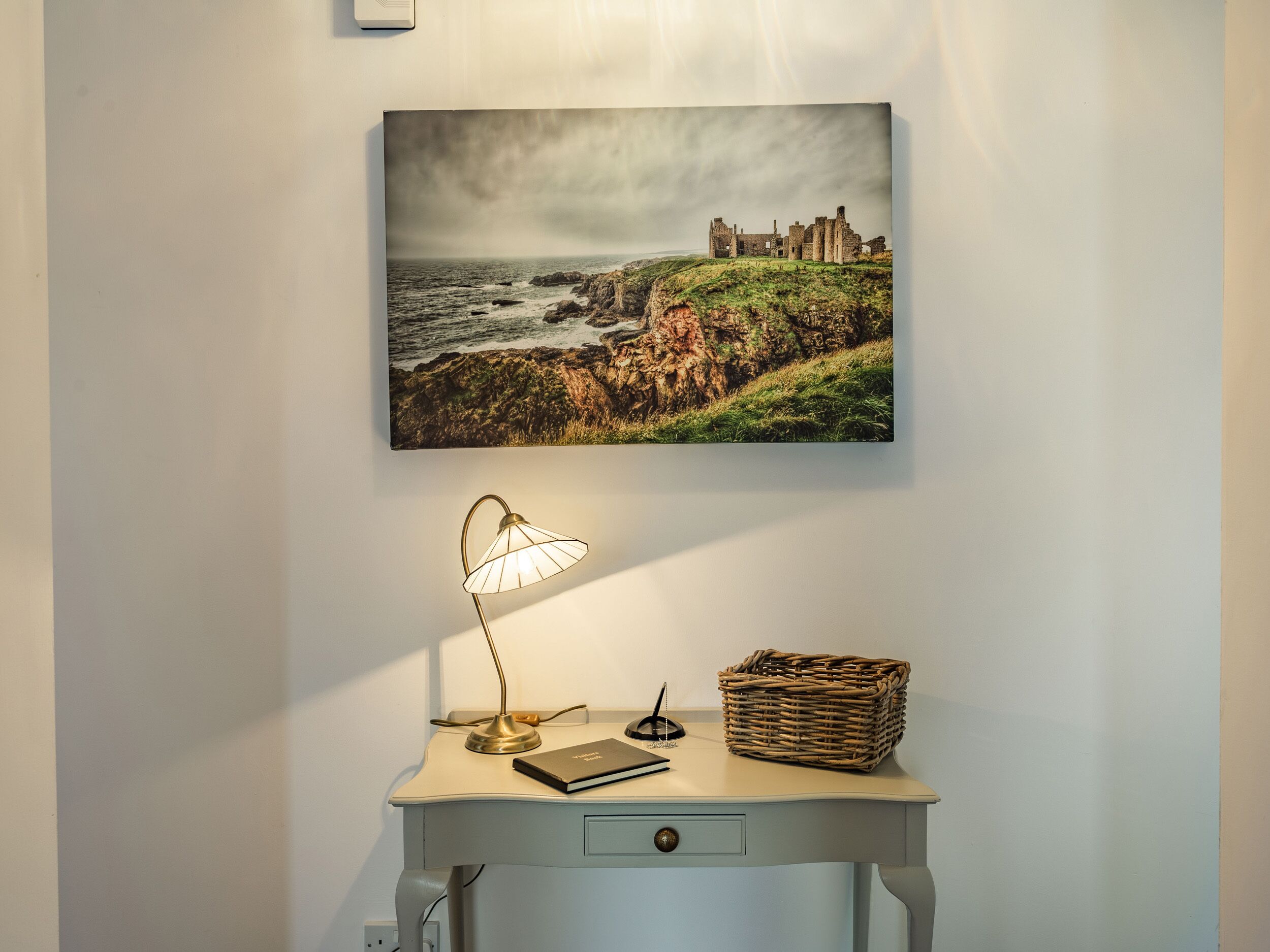 A desk with a lamp, a notebook, and a wicker basket against a wall with a framed picture of a coastal castle.