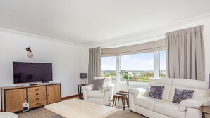 Ein modernes Wohnzimmer mit einem Flachbildfernseher, einer Holzkabine, einem beige gefärbten Sofa, einem Couchtisch und Blick auf einen Golfplatz durch große Fenster.