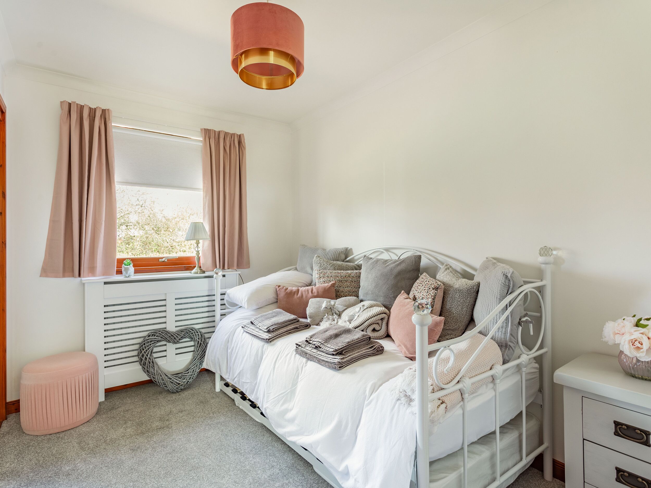 A bedroom with a white bed, pink curtains, a white radiator, a bedside table with a lamp, and a pink ottoman.