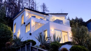Una casa moderna de dos pisos con balcón, rodeada de diversas plantas y árboles.