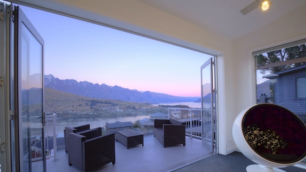 Habitación moderna con puerta de vidrio que da a un balcón con vistas a montañas y un lago. Hay sillones de mimbre y un sillón redondo acolchado en el balcón.