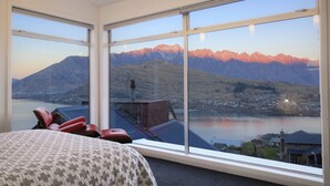 Habitación con amplios ventanales que ofrecen vistas a montañas y un lago. Hay sillones rojos y una cama con un edredón estampado.