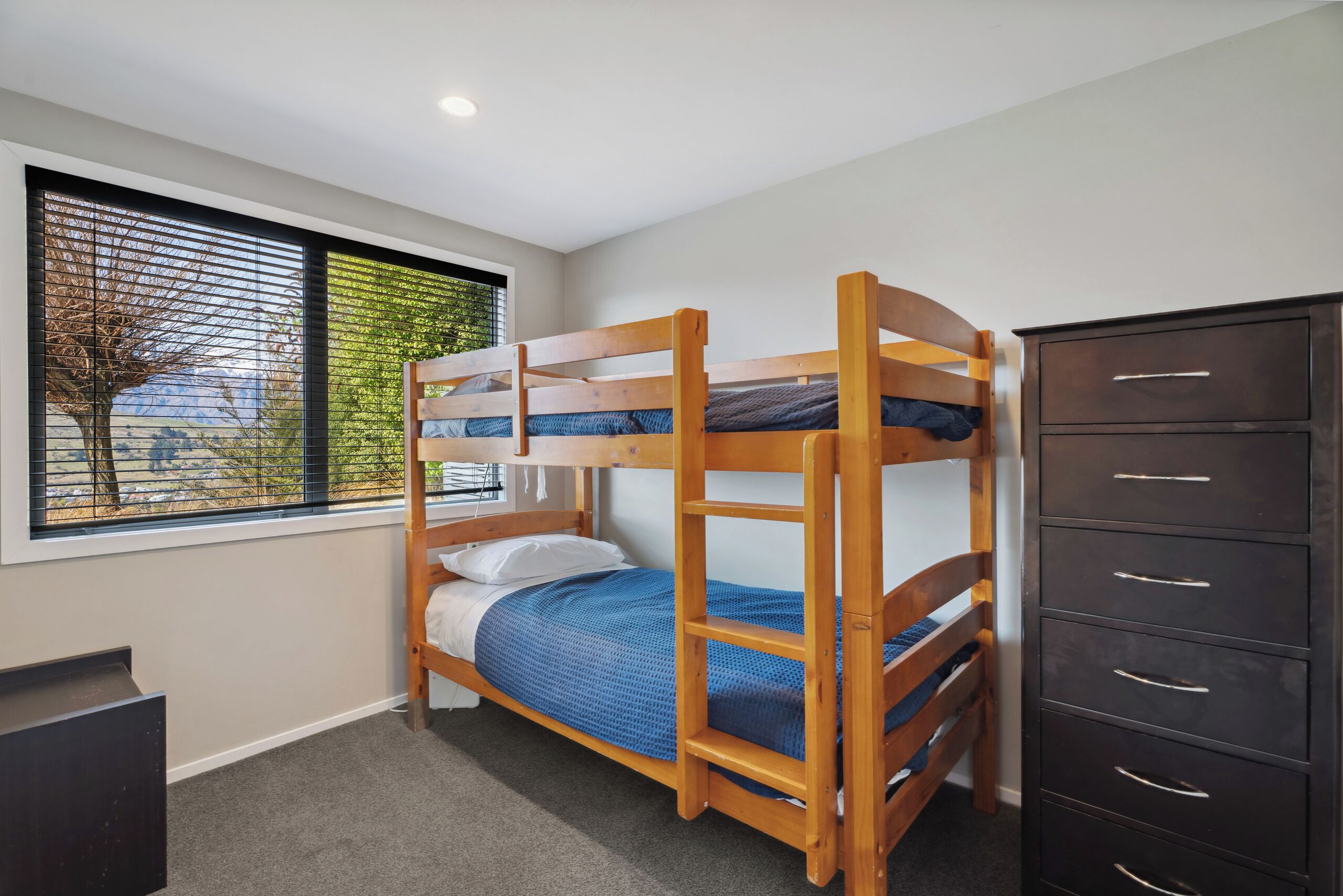 Una cama individual superpuesta con estructura de madera, un aparador con varios cajones y una ventana con persianas.