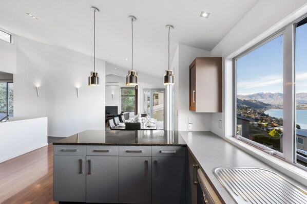 A modern kitchen with dark cabinets, a large island, pendant lights, and a view of a lake and mountains. - Paradise Peaks - Queenstown Holiday Home (Queenstown)