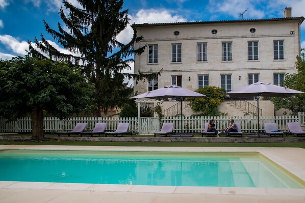 Un área de piscina con sillones de descanso y sombrillas, una cerca de piquetes blancos y un edificio grande con múltiples ventanas al fondo.