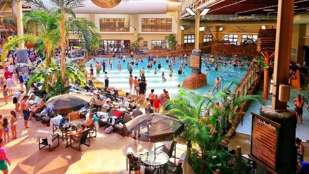 A large indoor swimming pool with multiple levels, surrounded by lounge areas with umbrellas and palm trees.