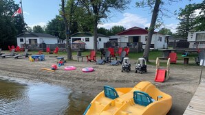 A sandy beach area with watercraft, beach chairs, and a wooden dock. - Chickadee #3 Graceland Cottages (West Nipissing)