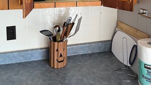 A kitchen with wooden cabinets, a shelf with glassware and plates, and a utensil holder with a jack-o'-lantern design. - Chickadee #3 Graceland Cottages (West Nipissing)