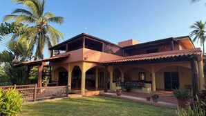 Une maison à deux étages avec un patio, entourée de palmiers et d’une pelouse bien entretenue.