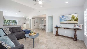 Amplia sala de estar con sofá, mesa de centro y mesa de comedor. Se observa un ventilador de techo, una cocina al fondo y un cuadro con motivos playeros en la pared.