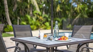 Una mesa con una botella, dos tazas y un plato de fruta en un entorno tropical.