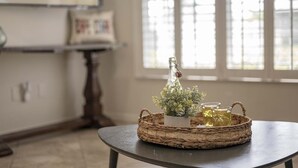 Una habitación con una mesa redonda, una cesta tejida con una botella y una planta, y una ventana con persianas.