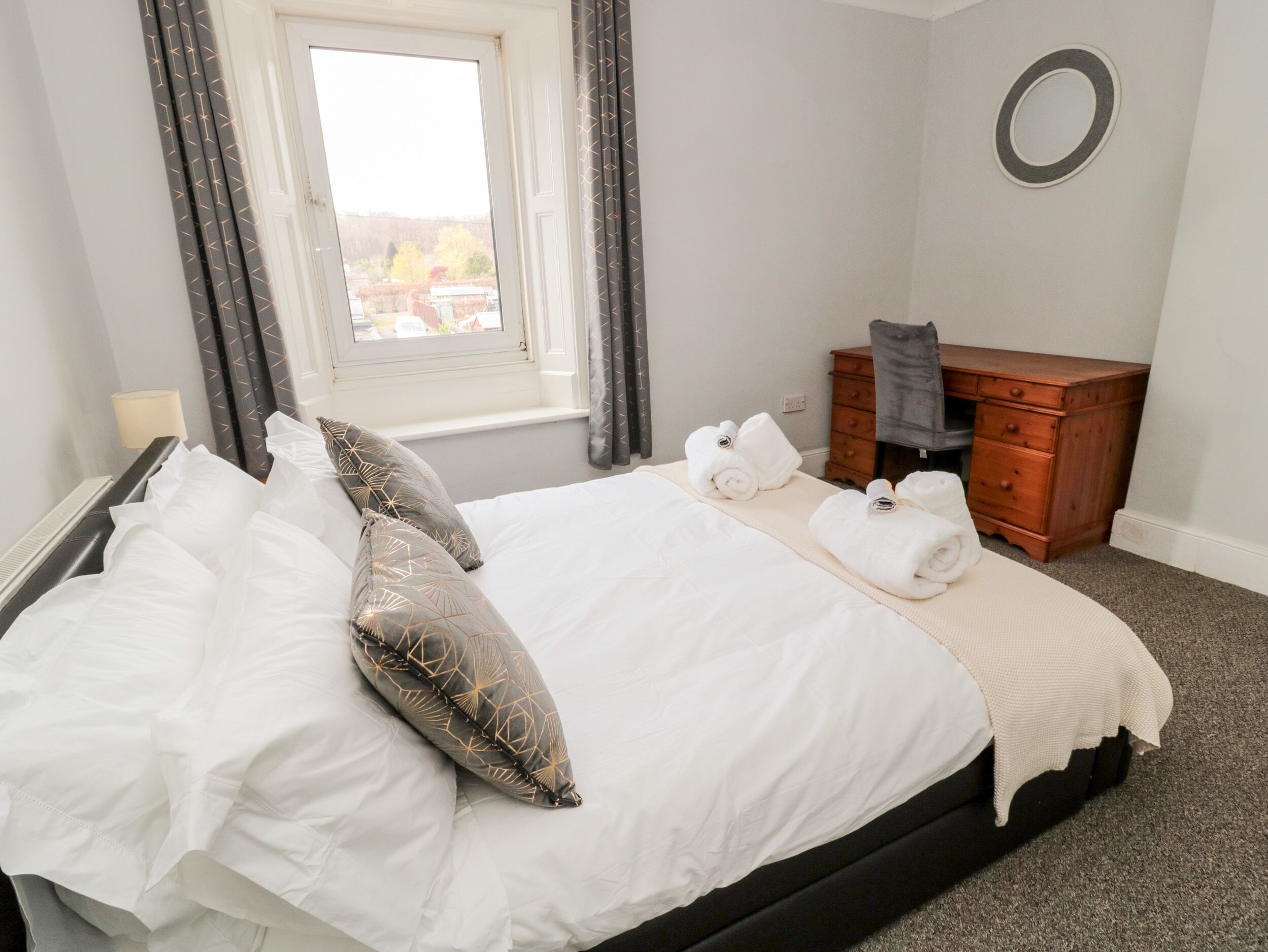A neatly made bed with white linens and decorative pillows, a wooden dresser, and a window with curtains.