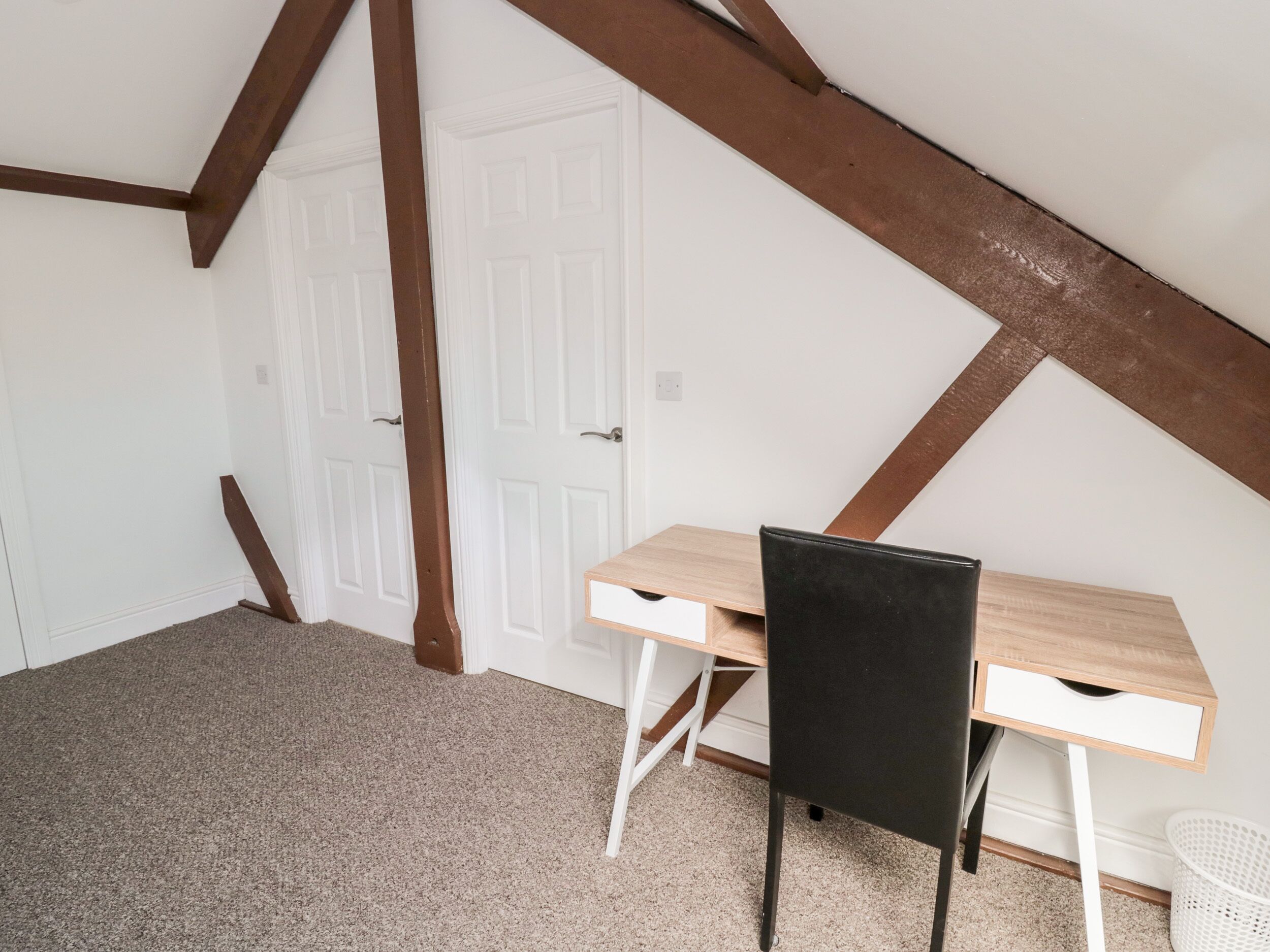 A room with a sloped ceiling, wooden beams, a desk, a chair, and a white door.
