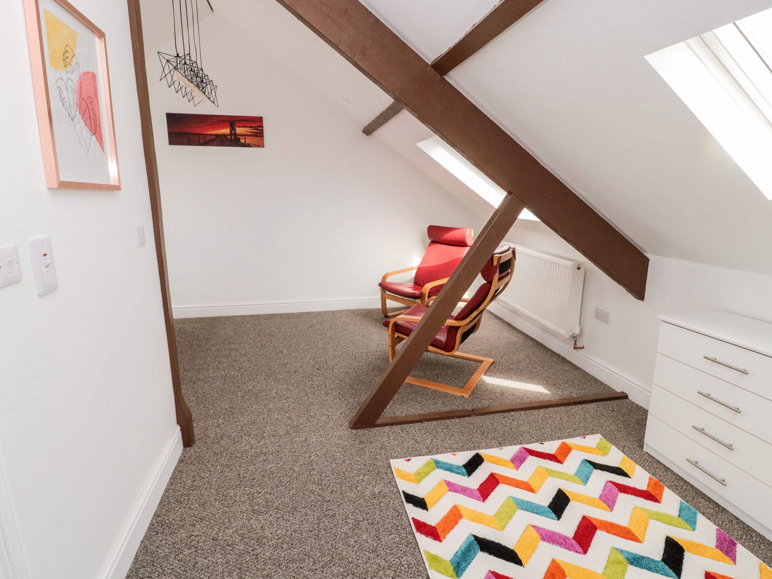 A room with a sloped ceiling, a red chair, a white dresser, and a colorful rug.