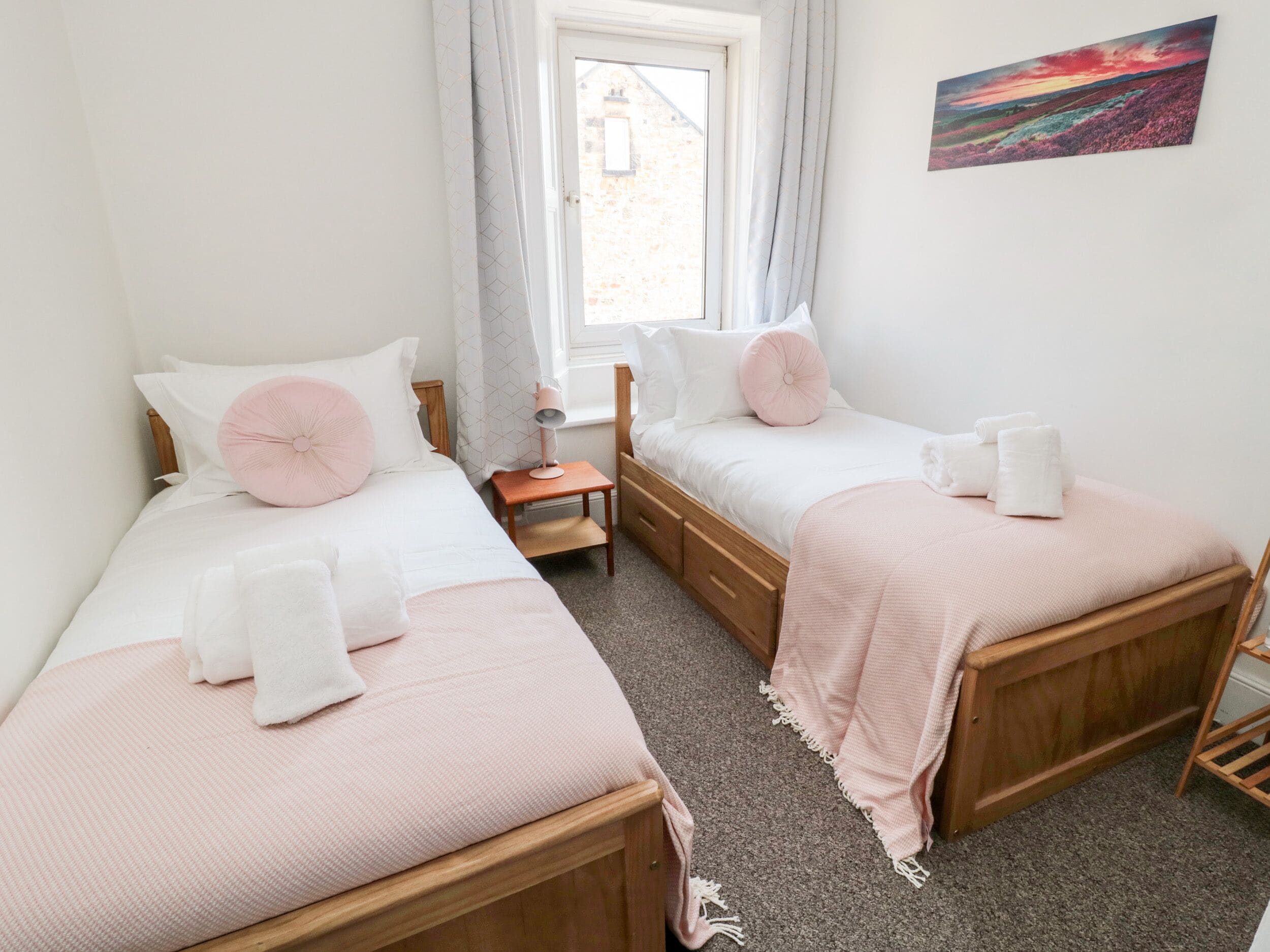 A bedroom with two single beds, a bedside table with a lamp, a window with curtains, and a painting on the wall.
