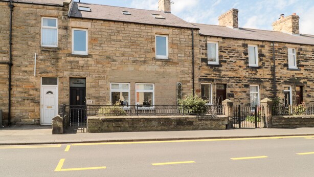 A row of stone terraced houses with numbered doors, a clear sky, and a road with yellow lines. - The Old Sweet Shop (Alnwick)