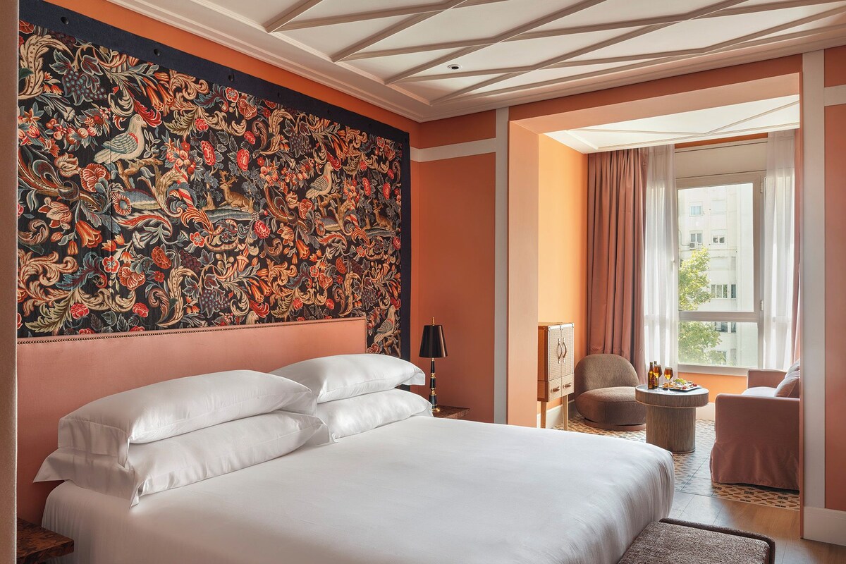 A hotel room with a large bed, a decorative wall tapestry, a seating area with a table, and a view of the outside through a window.