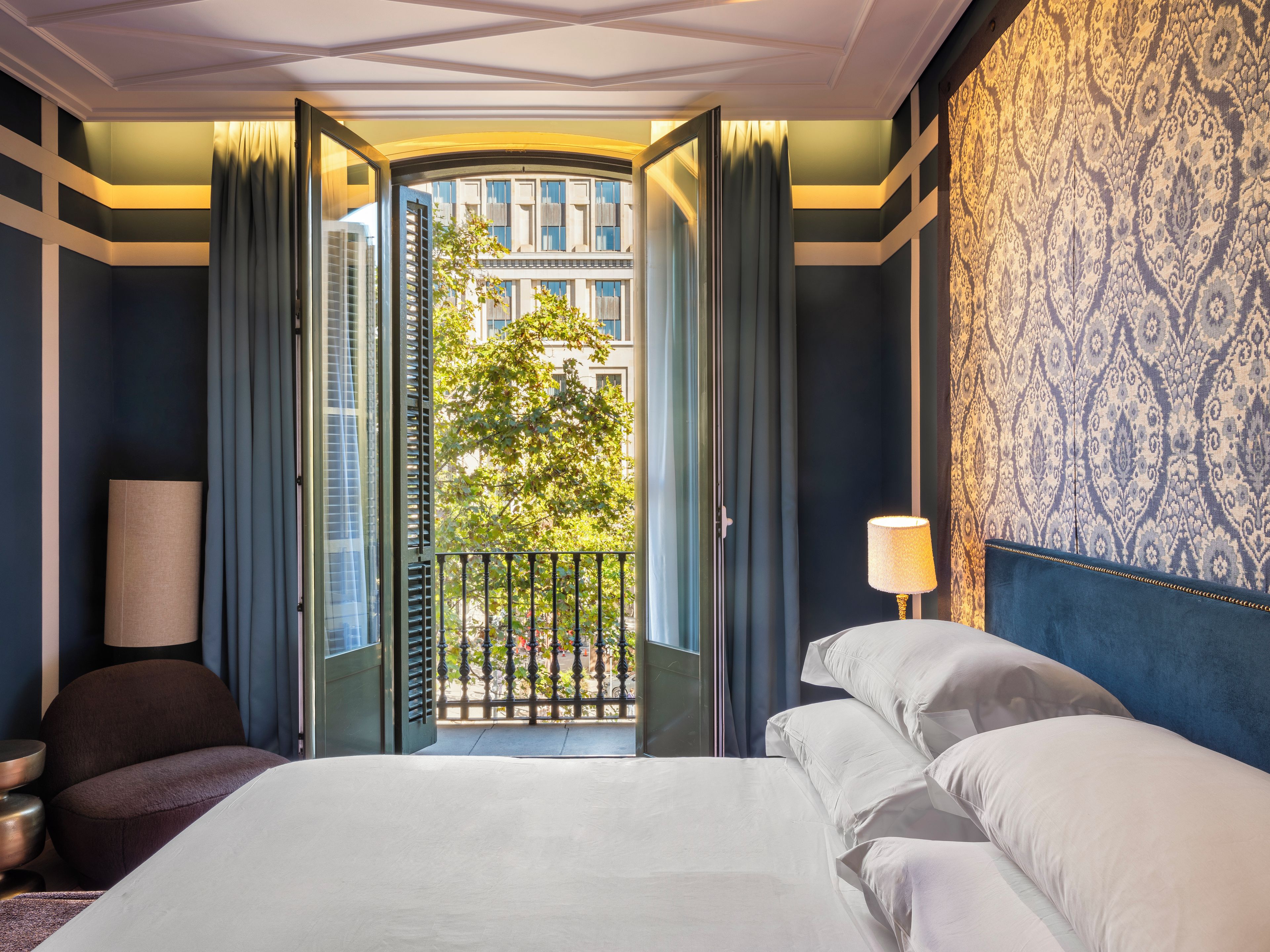 A hotel room with a bed, a nightstand, a lamp, and a view of a building through an open door.