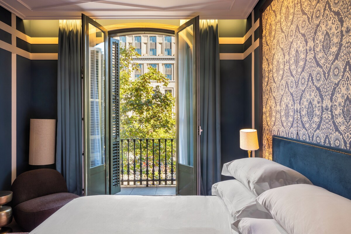 A hotel room with a bed, a nightstand, a lamp, and a view of a building through an open door.
