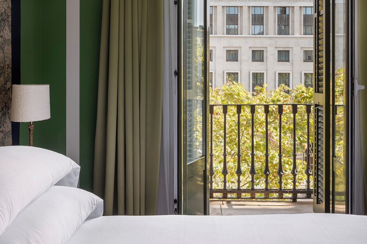 A hotel room with a bed, a window with curtains, and a view of a building outside.