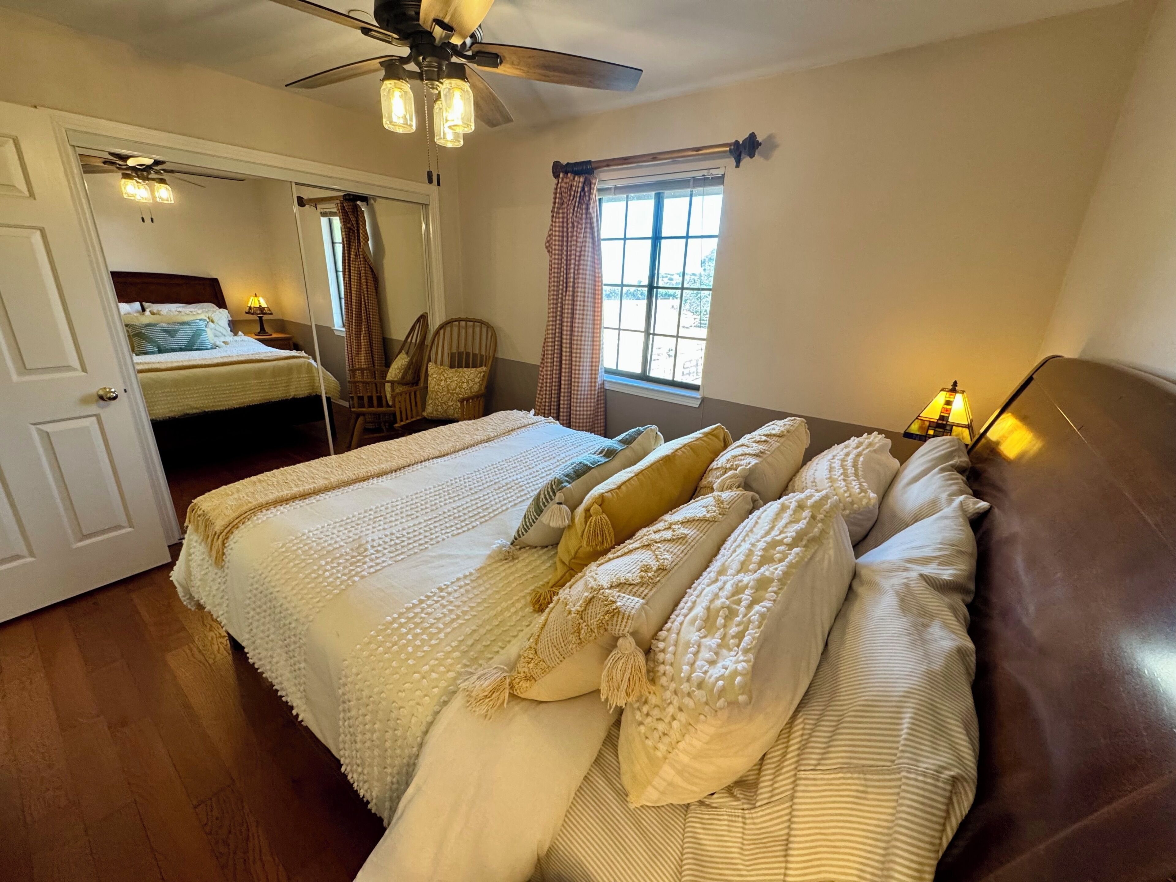 Een slaapkamer met een groot bed, een plafondventilator, een raam met gordijnen en een nachtkastje met een lamp.