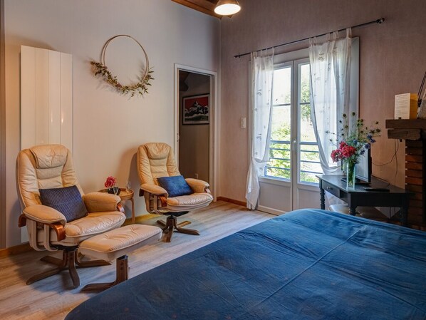 A room with two leather armchairs, a bed, a small table with a vase of flowers, and a view of greenery through the window.