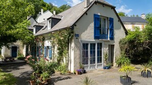 Un bâtiment pittoresque à la façade blanche et aux volets bleus, entouré de plantes en pot et bordé d’une allée gravillonnée.
