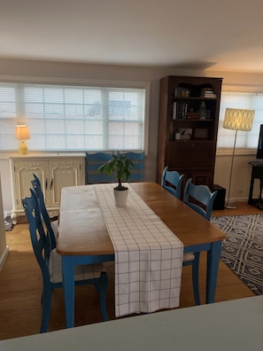 Un coin repas avec une table en bois, des chaises bleues, une plante en pot, un chemin de table blanc, une bibliothèque et une lampe.