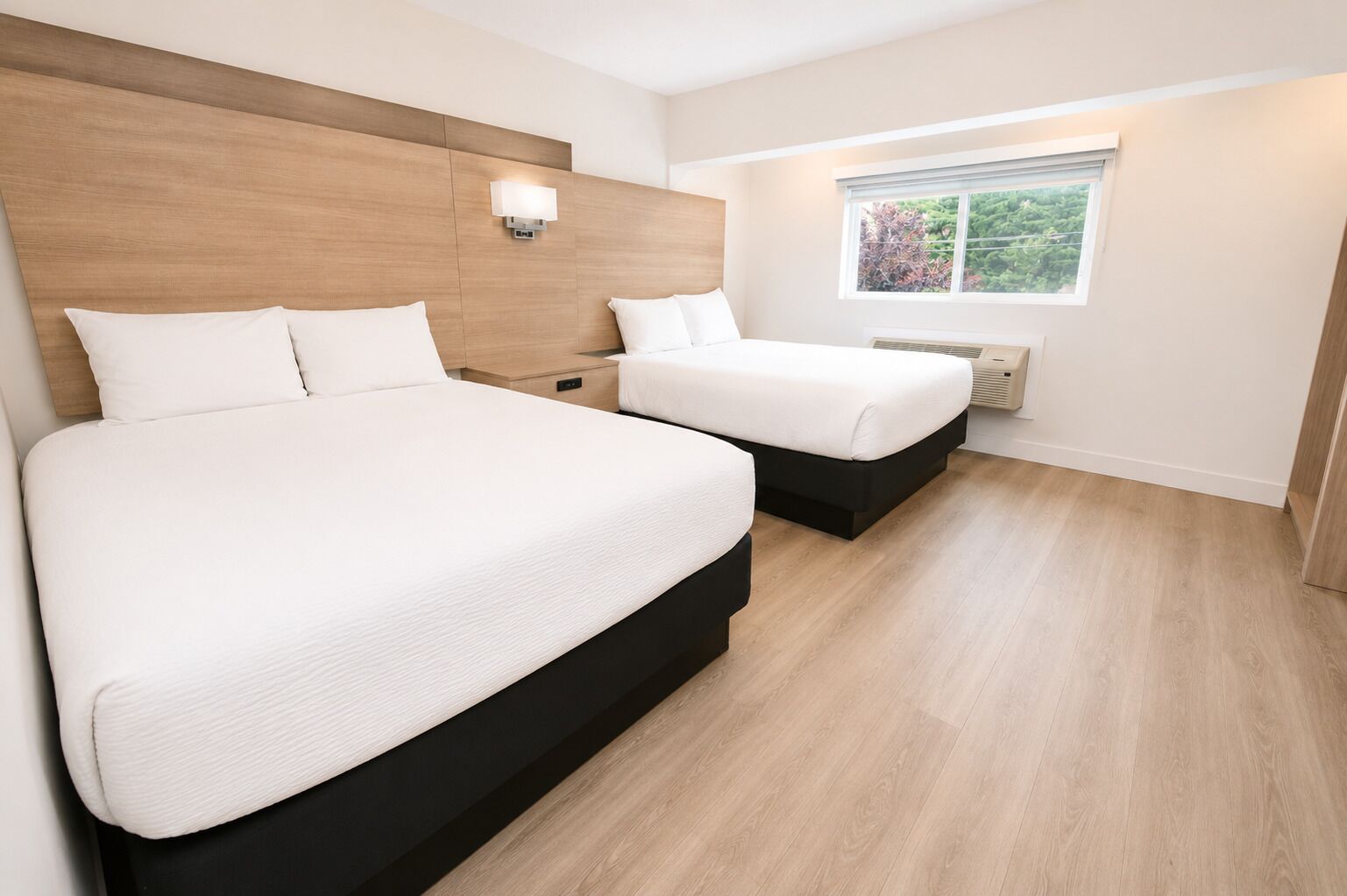 Habitación de hotel con dos camas, cabecera de madera, ventana con vista y lámpara fijada a la pared.