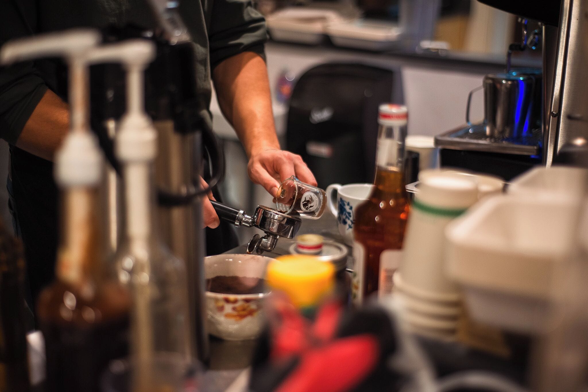 A person preparing a drink with a coffee machine, a bottle, and a mug on a counter.