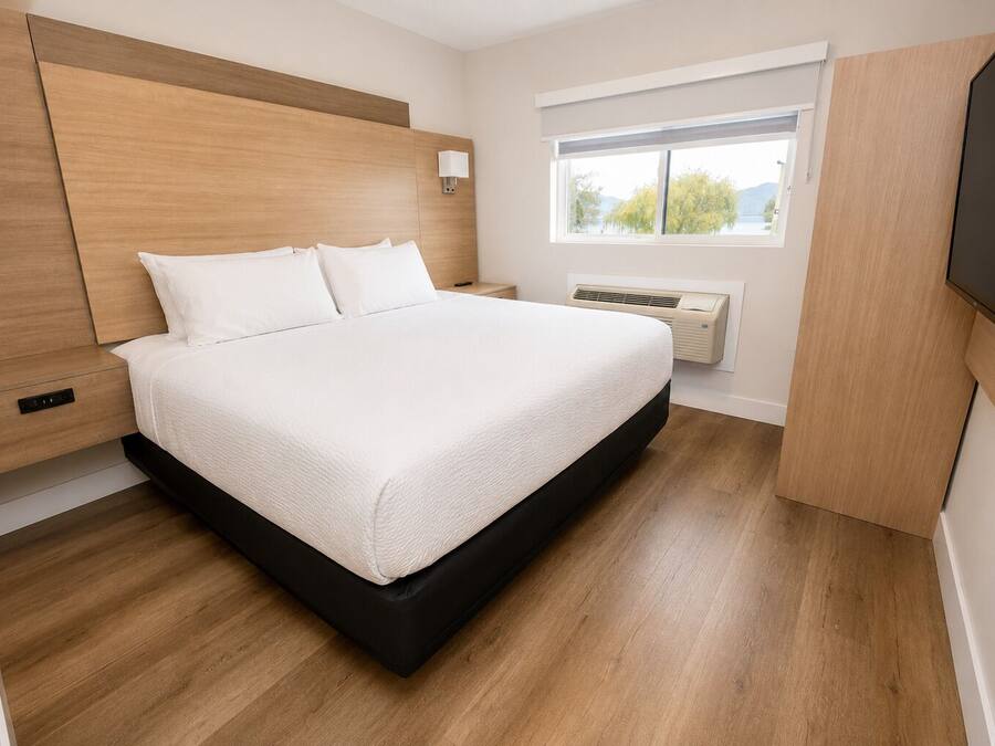 Ein modernes Schlafzimmer mit einem Holzkopfende, einem großen Bett mit weißer Bettwäsche, einem Nachttisch, einem an der Wand montierten Fernseher und einem Fenster mit Blick auf Bäume.