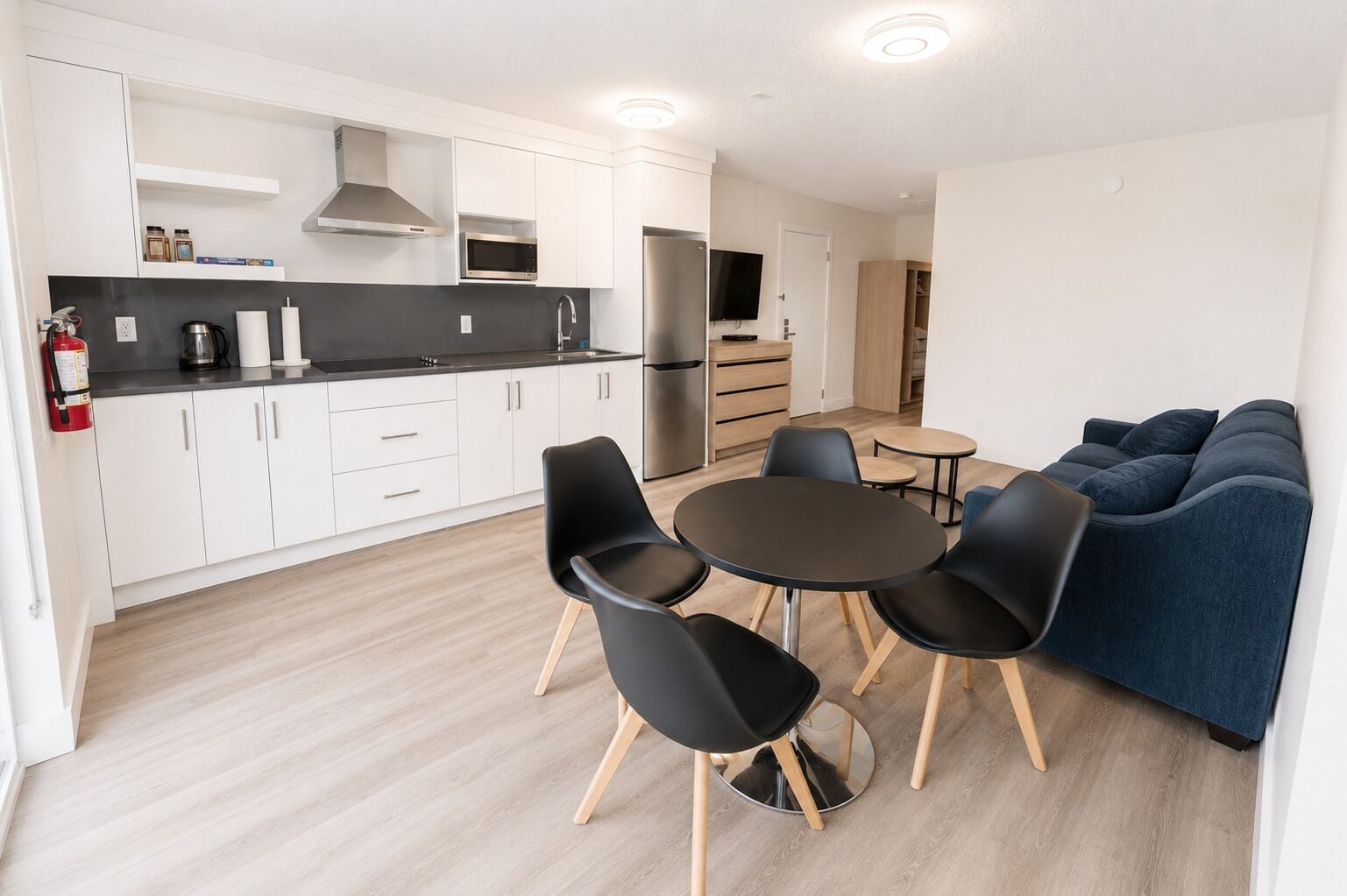 Una cocina moderna con armarios blancos, encimera negra y electrodomésticos de acero inoxidable. Hay un comedor con una mesa redonda y cuatro sillas, y un sofá azul en la sala de estar.
