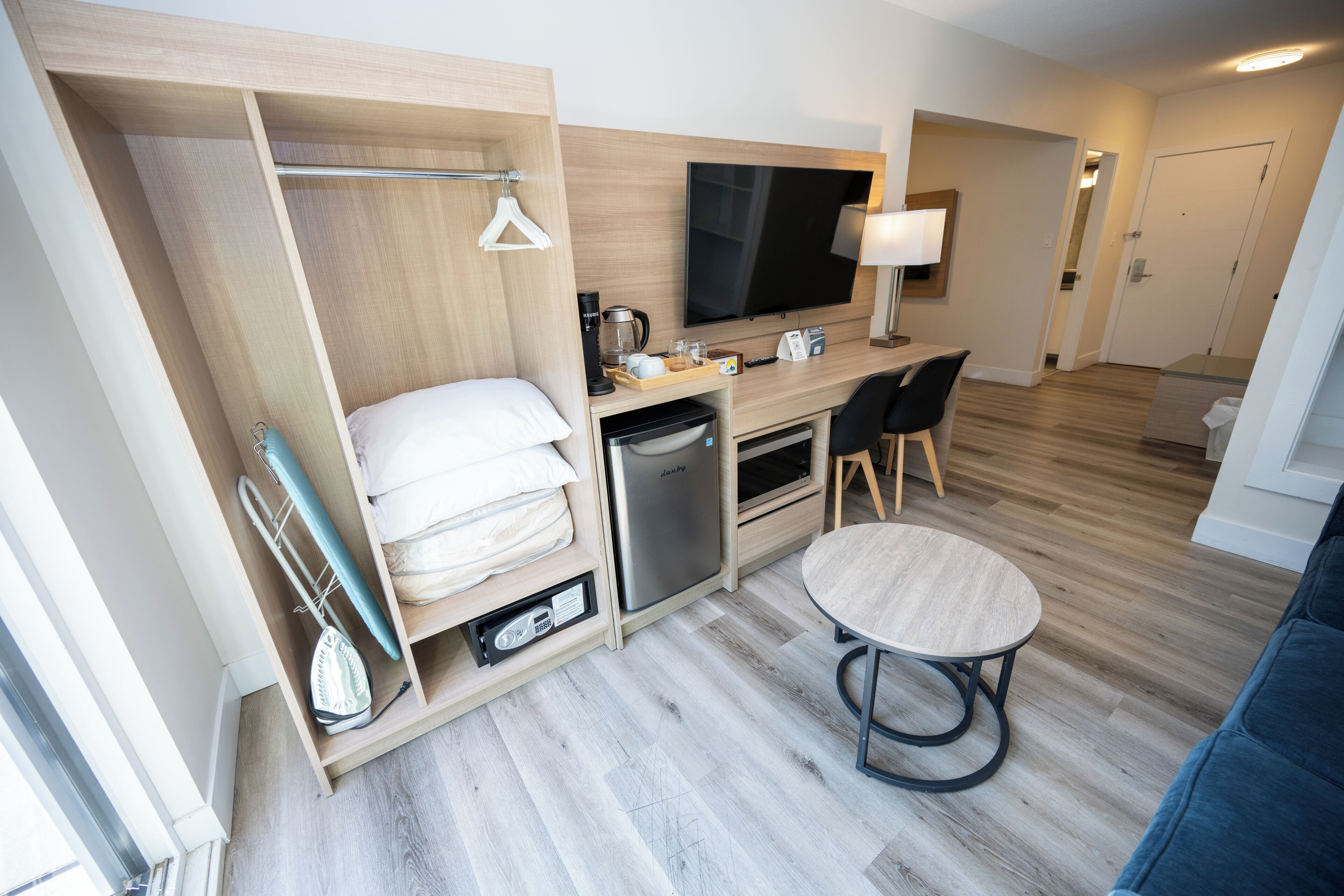 A compact hotel room with a bed, a desk with a TV, a coffee maker, and a small table.