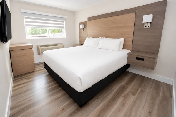 A modern bedroom with a large bed, bedside tables, a built-in heater, and a window with blinds. - Harrison Grand Motel LTD. (Harrison Hot Springs)