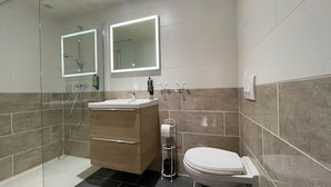 A modern bathroom with a glass-enclosed shower, a wooden vanity with a sink, a toilet, and two wall-mounted mirrors with illuminated frames. - Leclerc hôtel (Le Mans)