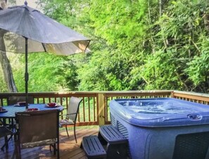 Relax on the deck with a hot tub, dining table, and comfortable seating under a shaded area surrounded by lush greenery.