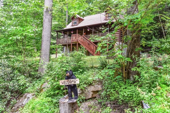 A rustic cabin surrounded by dense forest, with a wooden sign reading "WELCOME" on a tree stump.