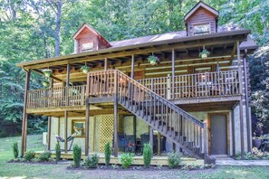 Step into this cozy cabin, featuring a spacious deck and a staircase leading to the upper level, with a large front door and a window overlooking the surrounding forest.
